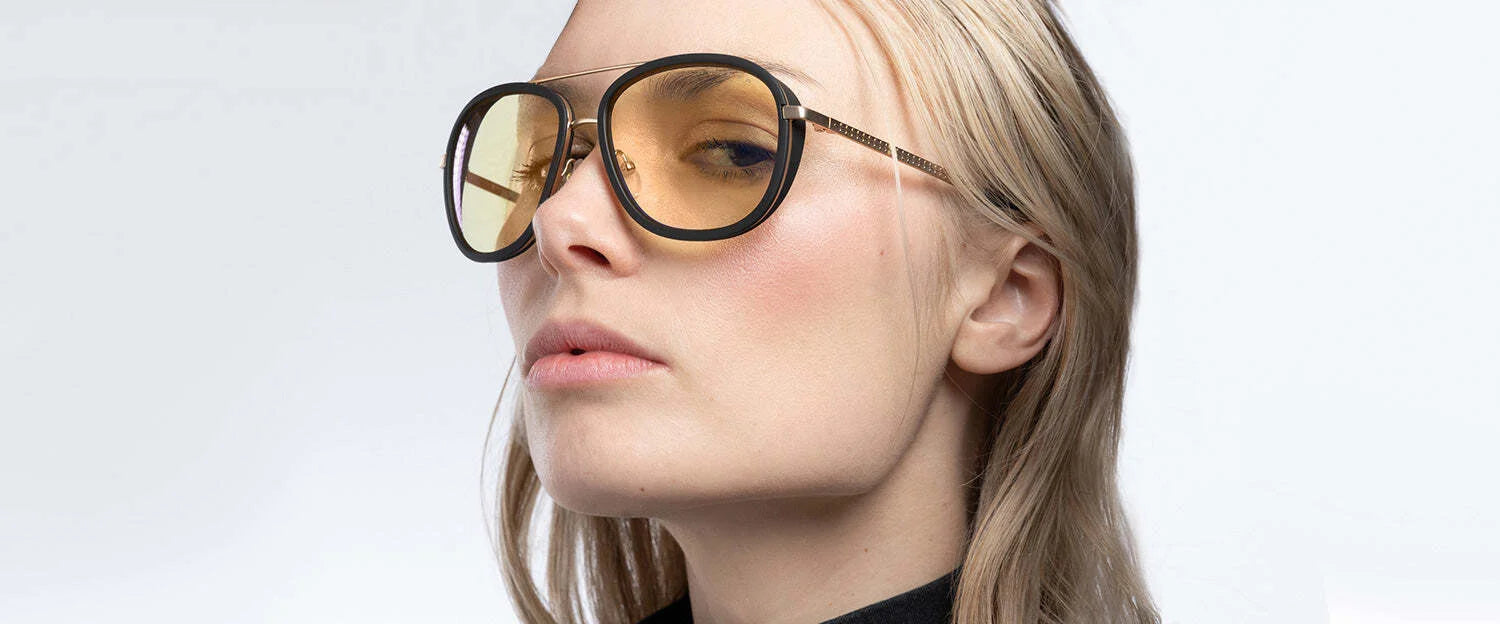 A person with long hair wears large, yellow-tinted Gunnar Dume Computer Glasses by Gunnar, featuring a stainless steel aviator frame and a black top, looking to the side.