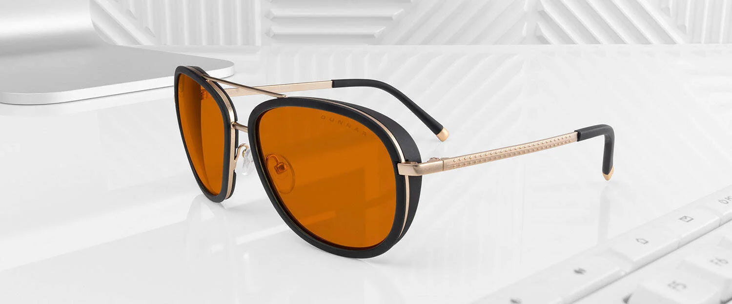 Gunnar Dume Computer Glasses, featuring stylish orange lenses and GUNNAR's blue light protection technology, rest on a white desk surrounded by a laptop and keyboard. With their sleek stainless steel aviator frame, they enhance any modern workspace.