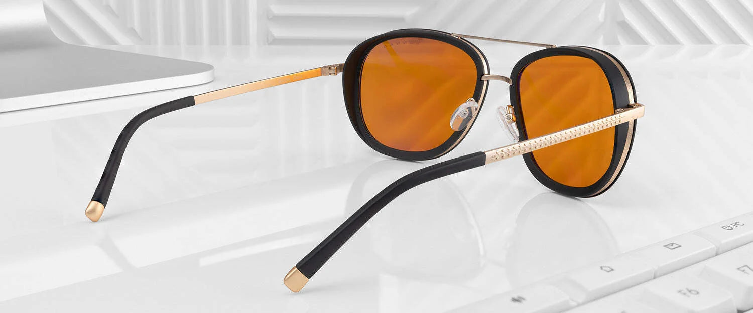 Gunnar Dume Computer Glasses, featuring black and gold frames with orange lenses, rest on a white reflective surface near a keyboard and laptop. Designed with a sleek stainless steel aviator frame, these glasses offer blue light protection perfect for extended screen time.