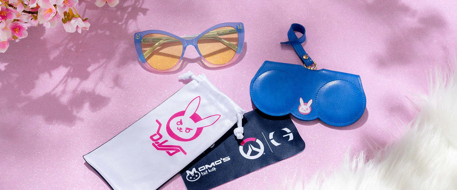The Gunnar Overwatch DVa Tokki Edition Computer Glasses rest elegantly on a flower-adorned pink surface with white fur. These size 54 glasses, featuring GUNNAR technology, come with a blue frame, matching case, and two branded pouches.