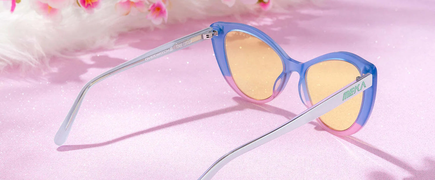 Gunnar Overwatch DVa Tokki Edition Computer Glasses with blue and pink frames and yellow lenses sit on a pink surface amid flowers. These stylish glasses capture the bold flair of the series, ideal for fans seeking vibrant style.