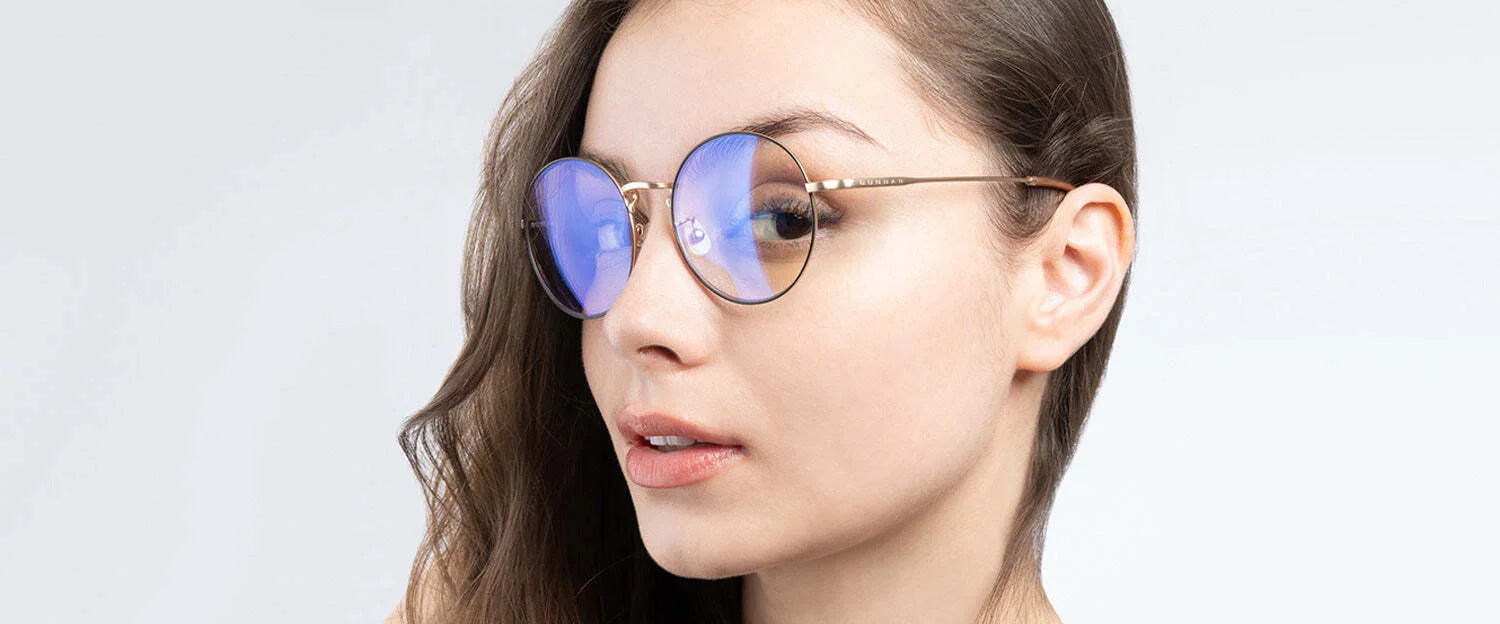 A young woman wearing Gunnar Ellipse Computer Glasses, featuring round blue-tinted lenses and a sleek stainless-steel frame, looks to the side against a plain background.