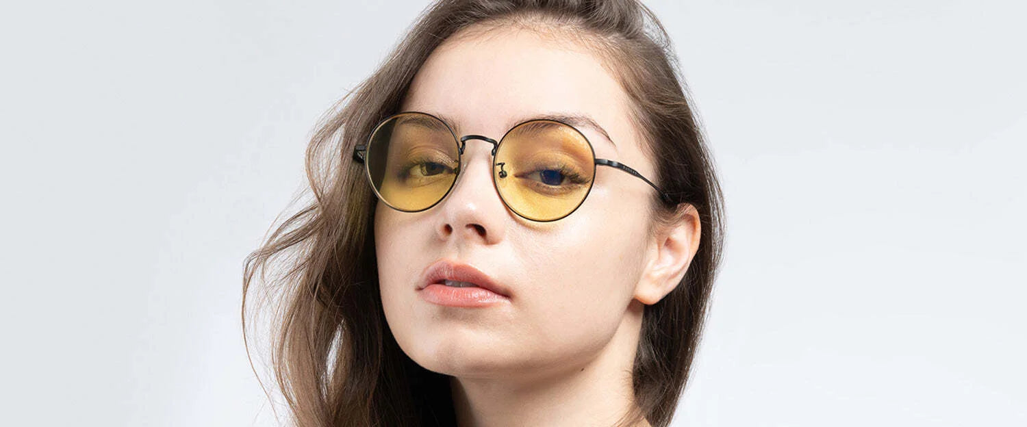 A person with long hair confidently looks at the camera, sporting Gunnar Ellipse Computer Glasses in size 51. The glasses feature a sleek stainless-steel frame and round, yellow-tinted lenses.