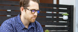 A man wearing Gunnar Emery Computer Glasses, featuring a premium frame design and anti-reflective lens coating, sits outdoors in front of a wooden fence. A small plant is in the background, adding to the serene setting.