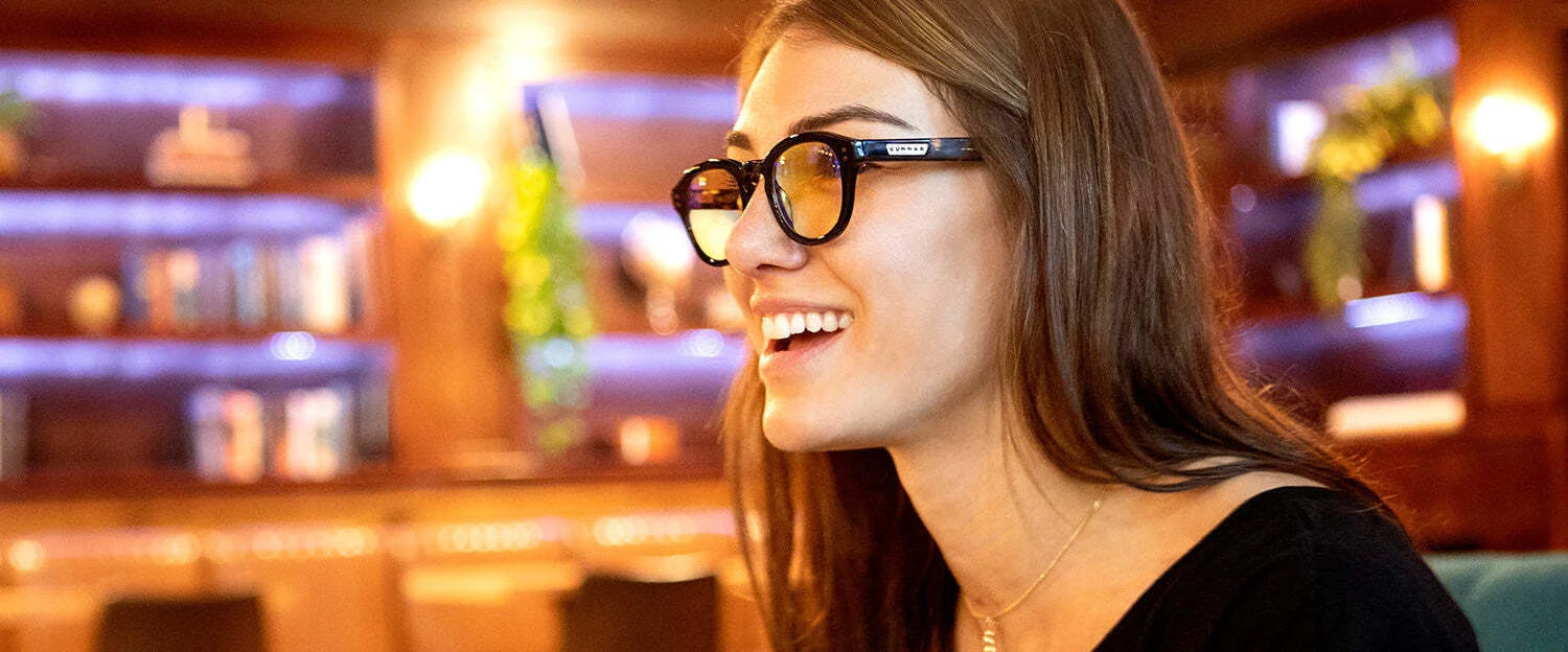 A woman wearing Gunnar Emery Computer Glasses in size 47, featuring an anti-reflective lens coating, smiles indoors against a blurred backdrop of shelves and lights.