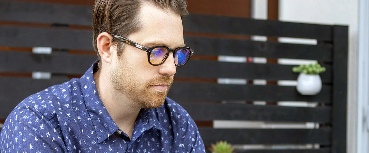 A man wearing Gunnar Emery Computer Glasses, featuring anti-reflective lenses, sits outdoors in a blue patterned shirt, focused. A wooden fence and plants are visible in the background.