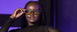 A person with braids wearing Gunnar Enigma Black Panther Edition Computer Glasses is in a black top, posing under purple lighting.