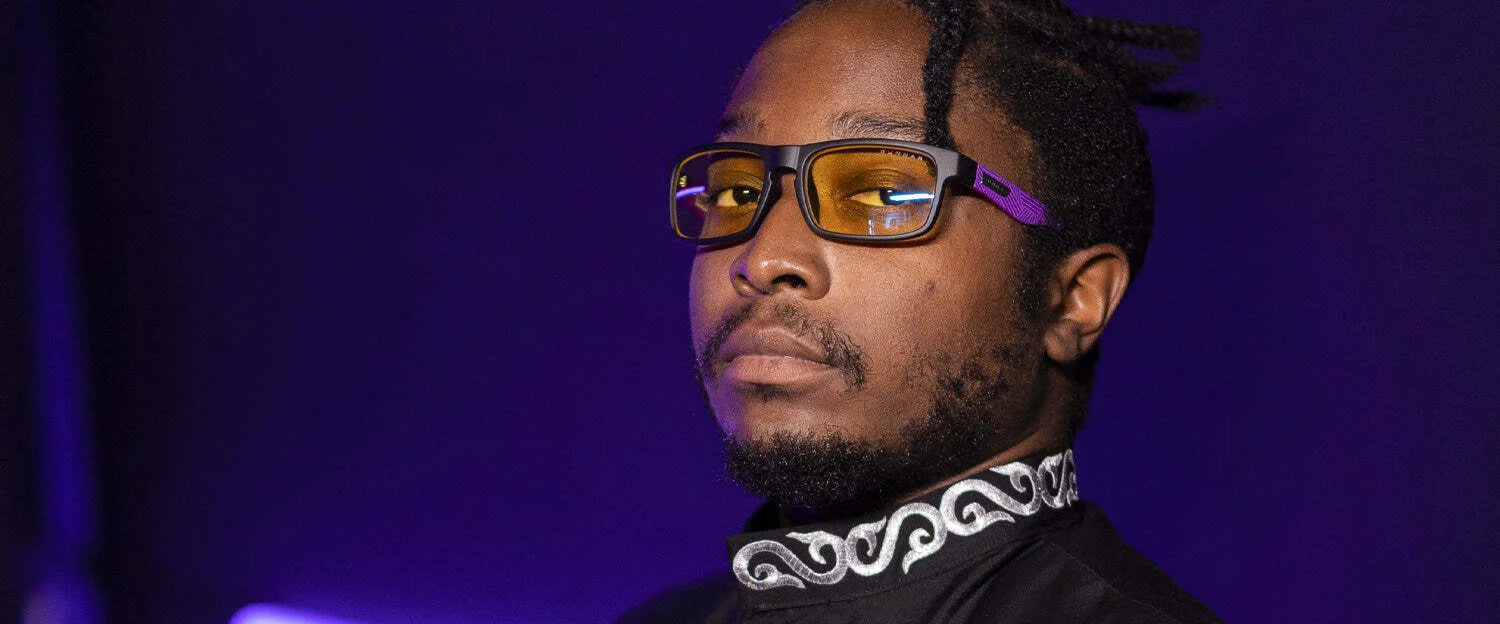 A man with braided hair wears Gunnar Enigma Black Panther Edition Computer Glasses, featuring patented lens technology. He gazes into the distance, dressed in a patterned shirt against a dark backdrop.