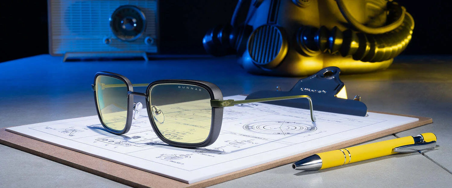 The Gunnar Fallout Vault 33 Computer Glasses in size 52, known for their retro-futuristic design and blue light blocking technology, rest on a clipboard with design sketches. A pen lies nearby, while a vintage radio and metal object create a scene blending classic and innovative elements.