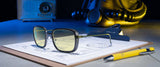 The Gunnar Fallout Vault 33 Computer Glasses rest on a clipboard with technical drawings, joined by a yellow pen, retro radio, and vintage helmet. Their design captures nostalgic charm and whispers stories of the past while hinting at future possibilities.