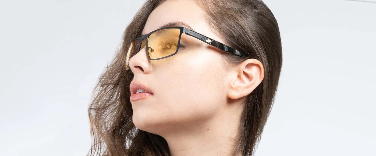 A woman with long hair wears Gunnar Fremont Computer Glasses, made of stainless steel and acetate, featuring yellow-tinted lenses that block blue light, as she gazes to the side against a plain background.