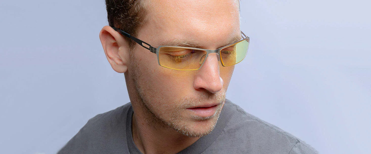 A man in a gray shirt, wearing yellow-tinted Gunnar Sheadog Computer Glasses against a light blue background, gazes down thoughtfully.