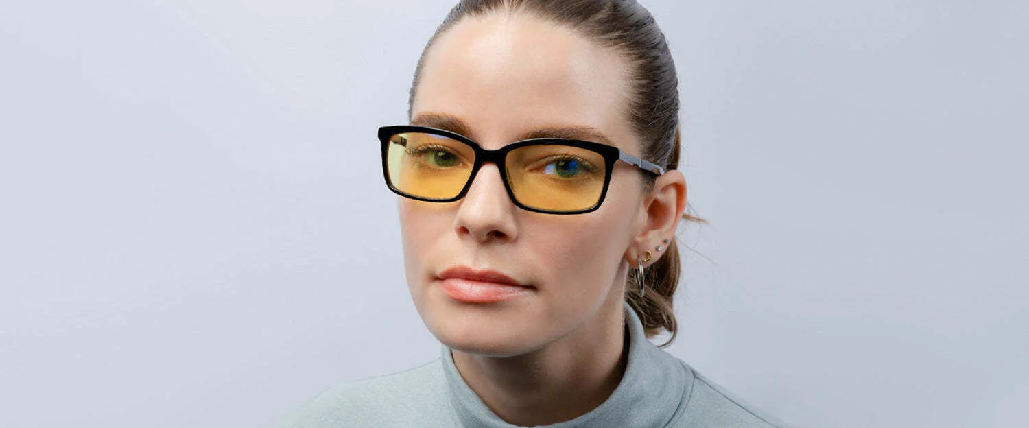 A woman wearing Gunnar Haus Computer Glasses by Gunnar with yellow-tinted lenses and a light gray turtleneck stands against a plain background.