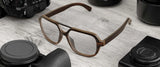 Gunnar Humboldt aviator-style ebony wood glasses sit elegantly on a wooden surface, amidst camera lenses and a sleek camera. Their distinctive design subtly incorporates Gunnar lens technology, enhancing both style and vision.