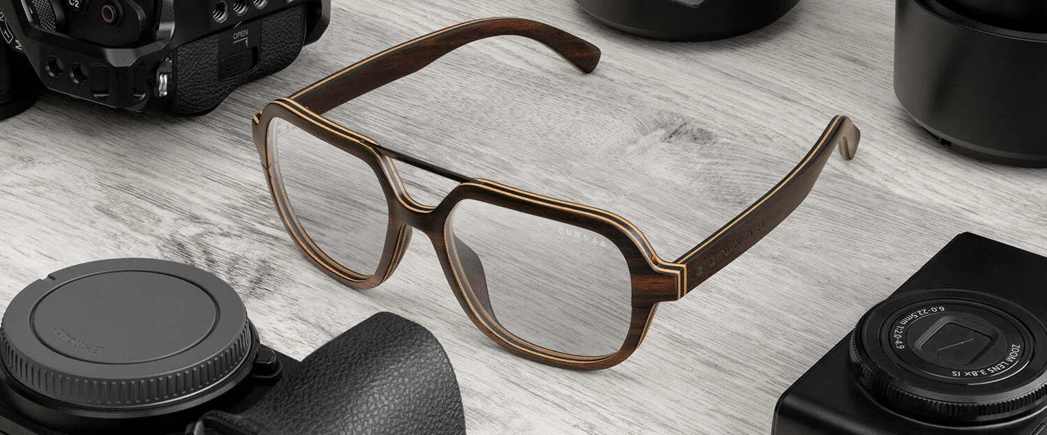Gunnar Humboldt aviator-style ebony wood glasses sit elegantly on a wooden surface, amidst camera lenses and a sleek camera. Their distinctive design subtly incorporates Gunnar lens technology, enhancing both style and vision.