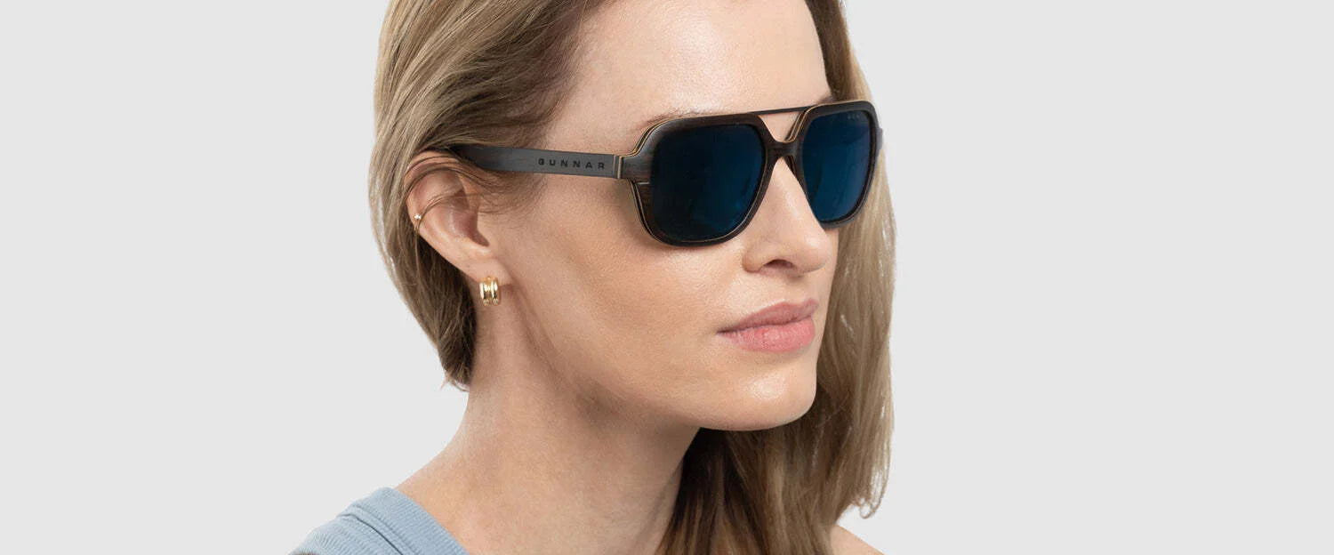 A woman wears Gunnar Humboldt Sunglasses with carbon fiber aviator-style frames that perfectly match her chic blue top against a stark white background.