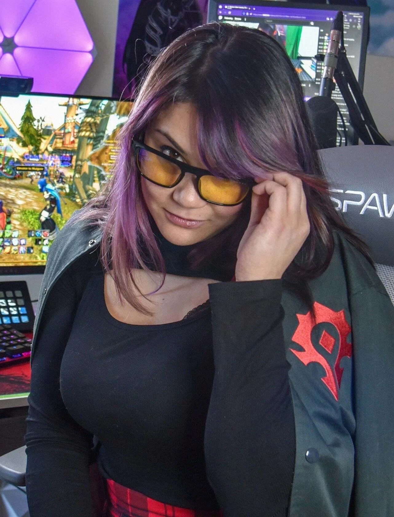 A person with purple-tinted hair and glasses, possibly Gunnar Intercept Computer Glasses for blue light protection, sits focused in a gaming setup with screens illuminating the background.