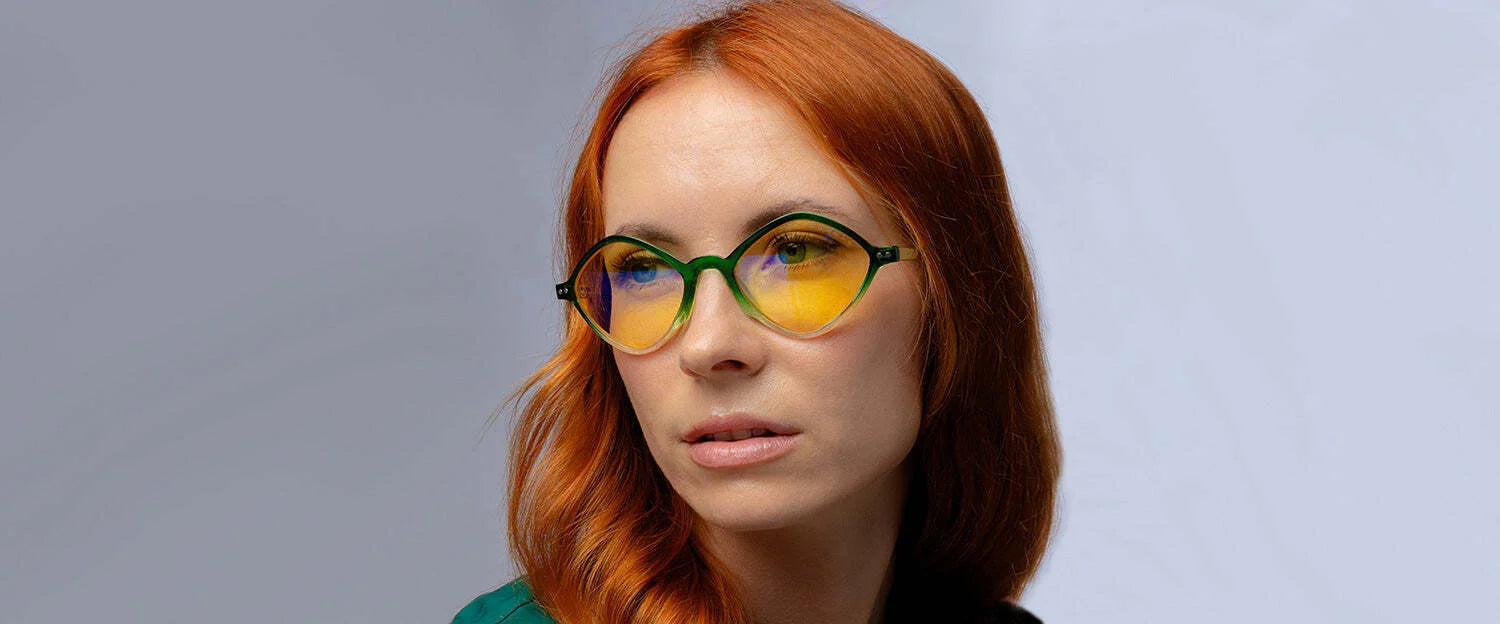 A red-haired individual dons Gunnar Loki Asgard Edition Computer Glasses | Size 53, with green round frames and yellow G-Shield™ Plus coated lenses, gazing sideways against a neutral backdrop.