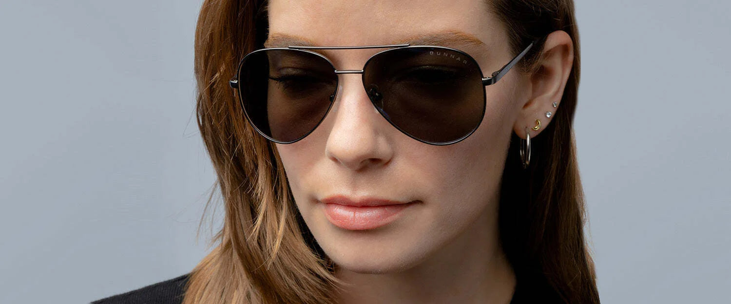 A person with long hair wears Gunnar Maverick Sunglasses size 58, which offer blue light protection, complemented by hoop earrings, while standing against a plain background.