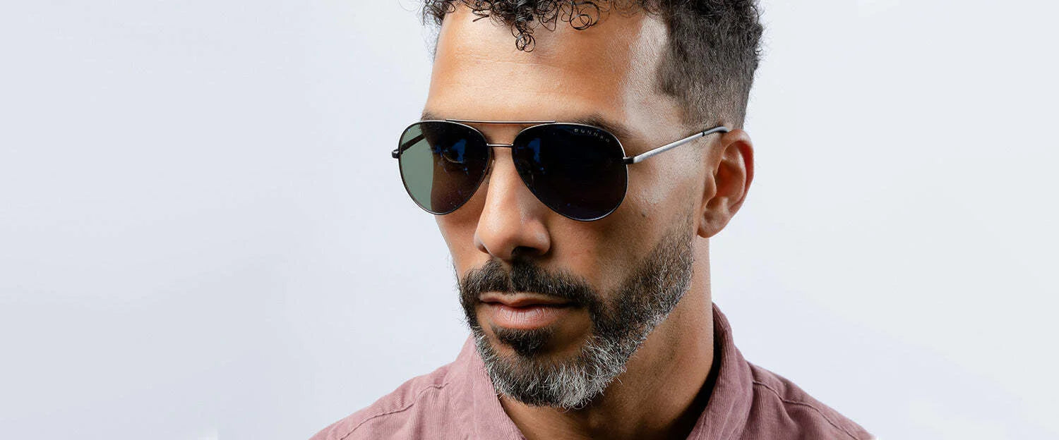 A man with curly hair and a beard sports Gunnar Maverick Sunglasses, featuring blue light protection, while wearing a mauve shirt against a light backdrop.