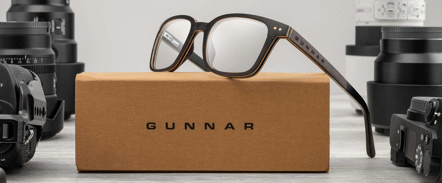 The Gunnar Muir Computer Glasses | Size 50 with sustainably sourced ebony wood black frames are displayed on a brown box labeled "Gunnar," surrounded by camera lenses and equipment.