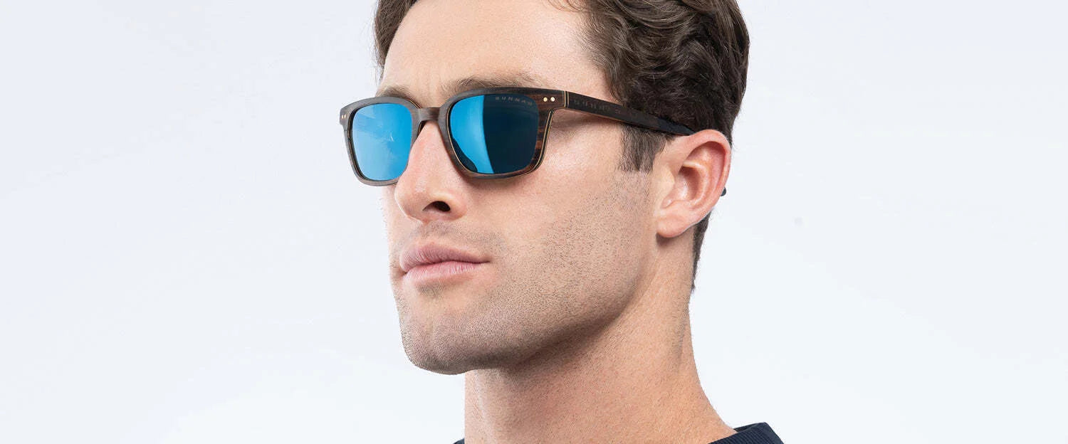 A person wearing Gunnar Muir Sunglasses with ebony wood frames and a navy shirt against a light background.