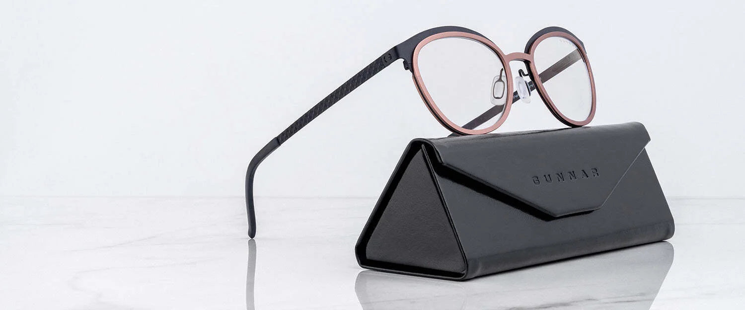 Gunnar Palomar Computer Glasses, featuring round black and pink frames, elegantly rest on a sleek closed black case. Positioned artistically on a pristine white marble surface, they showcase style with the added benefit of blue light blocking technology.