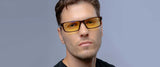 Sporting Gunnar Riot Computer Glasses with amber-tinted lenses for blue light protection, this person in a black shirt confidently looks at the camera against a gray background.