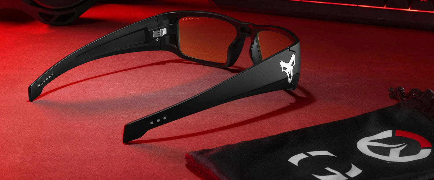 Gunnar Overwatch Reaper Computer Glasses with anti-reflective, orange lenses stylishly rest on a red surface next to the branded bag, all bathed in red ambient light—perfect for your next Overwatch 2 session.