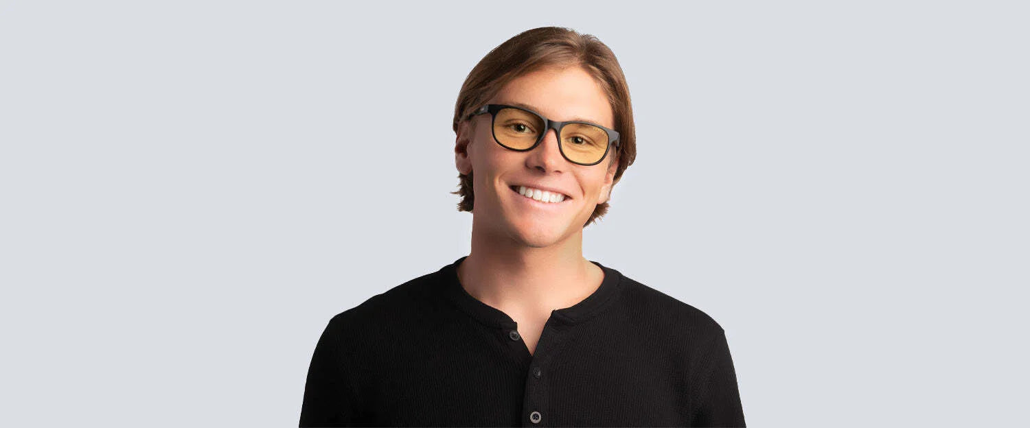 A smiling individual with glasses and brown hair wears a black shirt against a plain backdrop. Their Gunnar Rush Computer Glasses | Size 53 from Gunnar are crafted for comfort, efficiently blocking blue light.