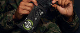 A person in camouflage, wearing Gunnar Call of Duty Tactical Edition Computer Glasses, carefully cleans the glasses with a black cloth adorned with a green laurel and skull graphic.