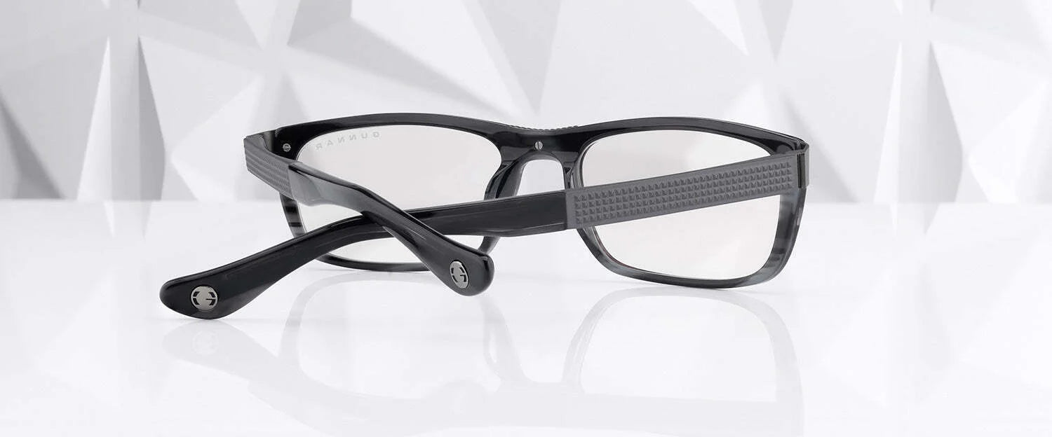 Gunnar Tahoe Computer Glasses (Size 55) by Gunnar elegantly rest on a glossy white surface with a geometric white backdrop. Featuring a black frame with textured arms, these glasses combine style and blue light blocking technology for eye protection.