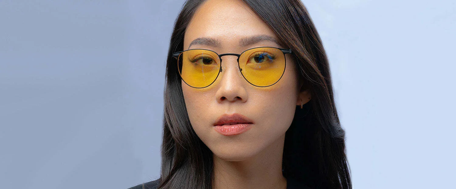 Wearing Gunnar Mateo Computer Glasses, the person flaunts yellow-tinted lenses with GUNNAR's technology in sleek black. These stylish specs, set against a light blue backdrop, block blue light effectively and rest on a lightweight titanium frame for comfort.