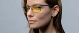 A long-haired individual with multiple earrings dons Gunnar Mendocino Computer Glasses, featuring a lightweight titanium frame and yellow lenses designed for blue light blocking, set against a gray background.