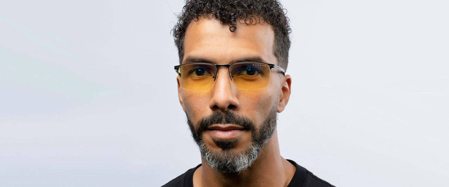 A curly-haired man with a beard wears Gunnar Marin Computer Glasses, Size 54. These blue light blocking glasses feature a sleek titanium frame and yellow-tinted lenses, set against a plain background.