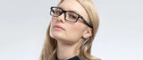 A person with long blonde hair dons stylish Gunnar Vertex Computer Glasses, which offer blue light protection and feature a nylon frame, while wearing a sleek black top. They gaze upwards against a light background, highlighting the eyewear's durability.