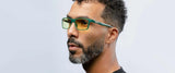 A bearded man with curly hair is wearing Gunnar Vertex Computer Glasses, Size 55, featuring durable nylon frames with green accents and yellow lenses for stylish blue light protection.