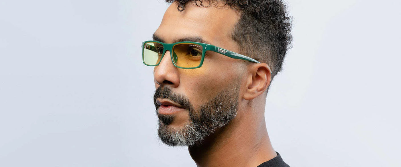 A man with curly hair and a beard is sporting Gunnar Vertex Computer Glasses | Size 55 in a green-yellow shade, designed by Gunnar for blue light and UV protection. He pairs them with a black shirt against a light background.