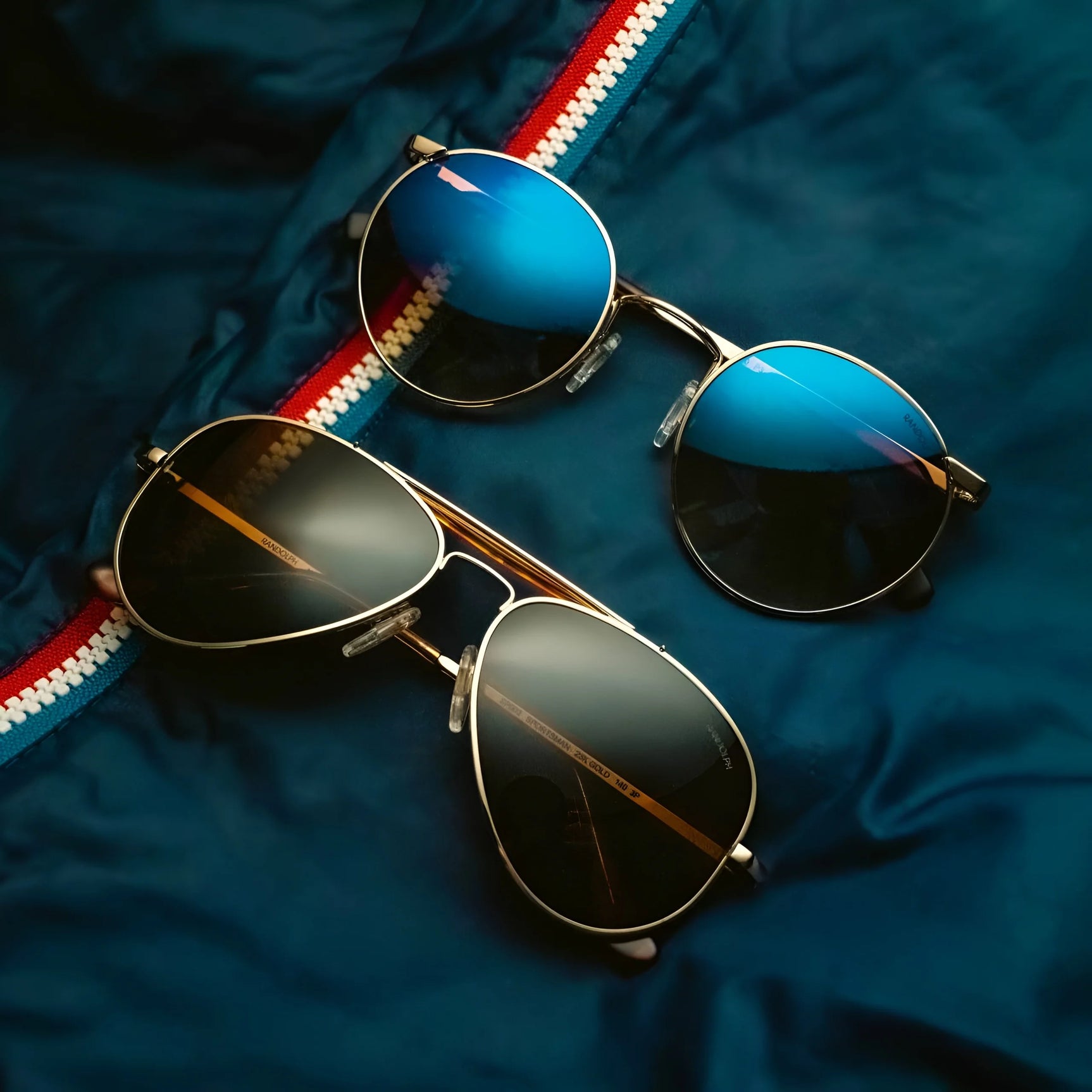 Two pairs of Randolph SPORTSMAN Sunglasses featuring blue and black lenses are placed on dark blue fabric accented with a striped edge, perfect for your outdoor adventures while delivering crucial UV protection.