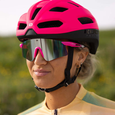 A person wearing Rudy Project Astral Sunglasses (size 147) and a bright pink bike helmet stands outdoors, greenery in the background.