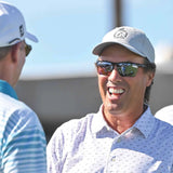 A man in a white golf shirt and gray cap, wearing Scheyden CIA GRABBER Sunglasses with LT33 RSR lenses, smiles while talking to another man in a blue and white striped shirt.