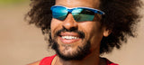 Grinning widely, the person wears a red tank top and blue Tifosi Optics VERO Sunglasses, Size 64, perfect for smaller faces. With a beautiful beach reflected in the lenses, these sunglasses excel in style and performance for multi-sport activities.