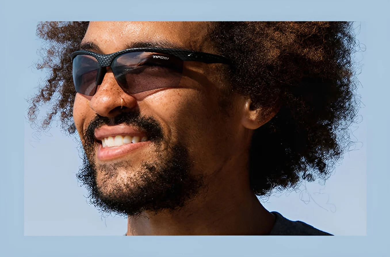 A person with curly hair and Tifosi Optics VERO Sunglasses smiles against a blue sky. Perfect for smaller faces, these sunglasses blend style and functionality, making them ideal for multi-sport activities.