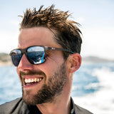 A man with wet hair and beard wears Julbo Creek Sunglasses, featuring a lightweight frame and Spectron HD lenses, smiling on a boat as the ocean and sky reflect beautifully.