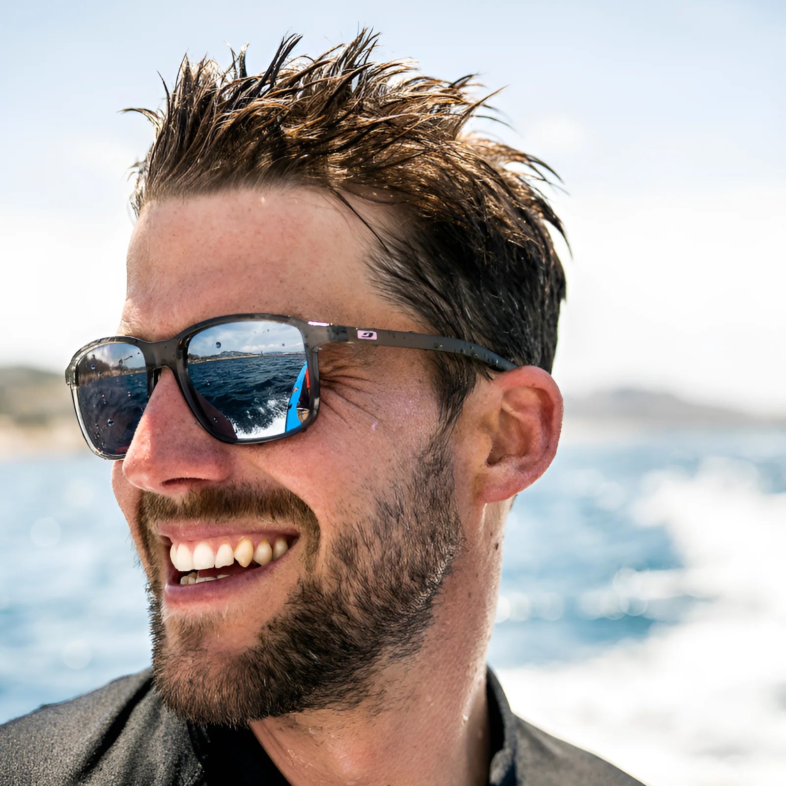 A man with wet hair and beard wears Julbo Creek Sunglasses, featuring a lightweight frame and Spectron HD lenses, smiling on a boat as the ocean and sky reflect beautifully.