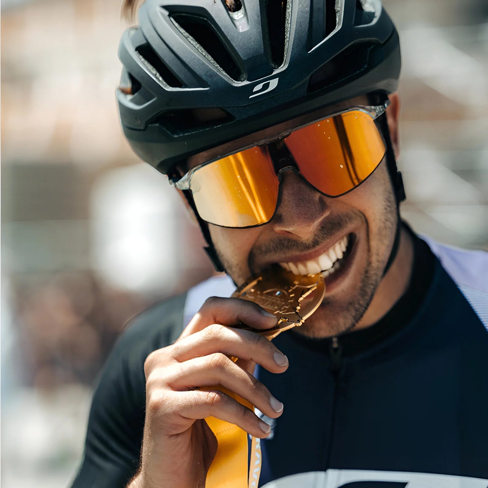 A cyclist wearing a helmet and Julbo Density Sunglasses with CustomFit Temples bites a gold medal while smiling.