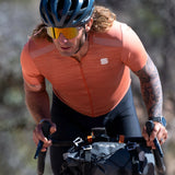 A long-haired cyclist rides uphill wearing an orange shirt, helmet, and Julbo Density Sunglasses with CustomFit Temples for comfort; the eyewear shields his eyes while bags sit on the bike.