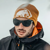 Wearing a black jacket with orange lining, a man in an orange beanie and sunglasses confidently stands in a snowy setting. His Julbo Explorer 2.0 Sunglasses Size 61 shield his eyes from the harsh alpine environment with high-protection lenses.
