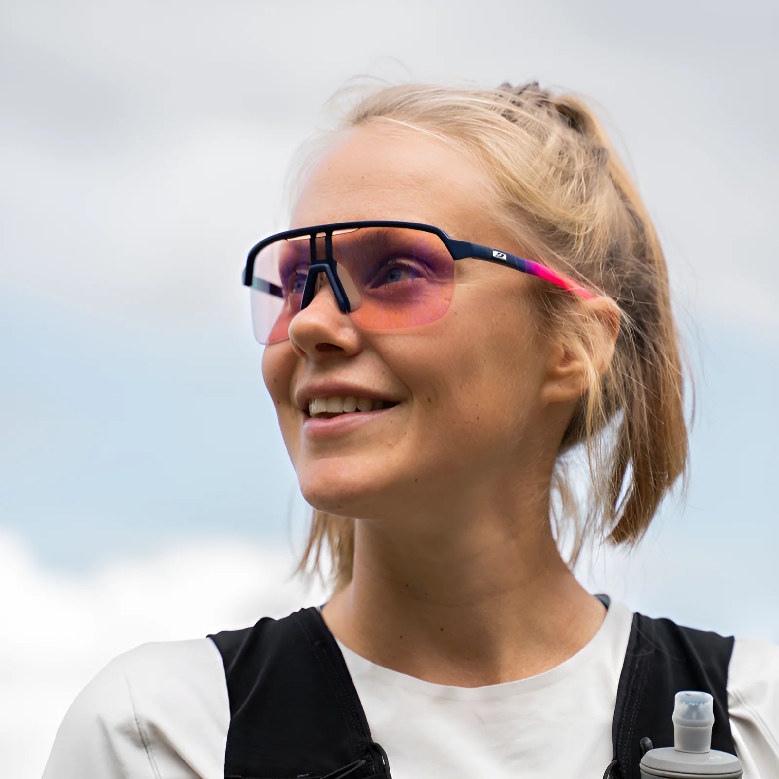 A woman outdoors, smiling under a partly cloudy sky in her Julbo Frequency Sunglasses, is ready for her next adventure.