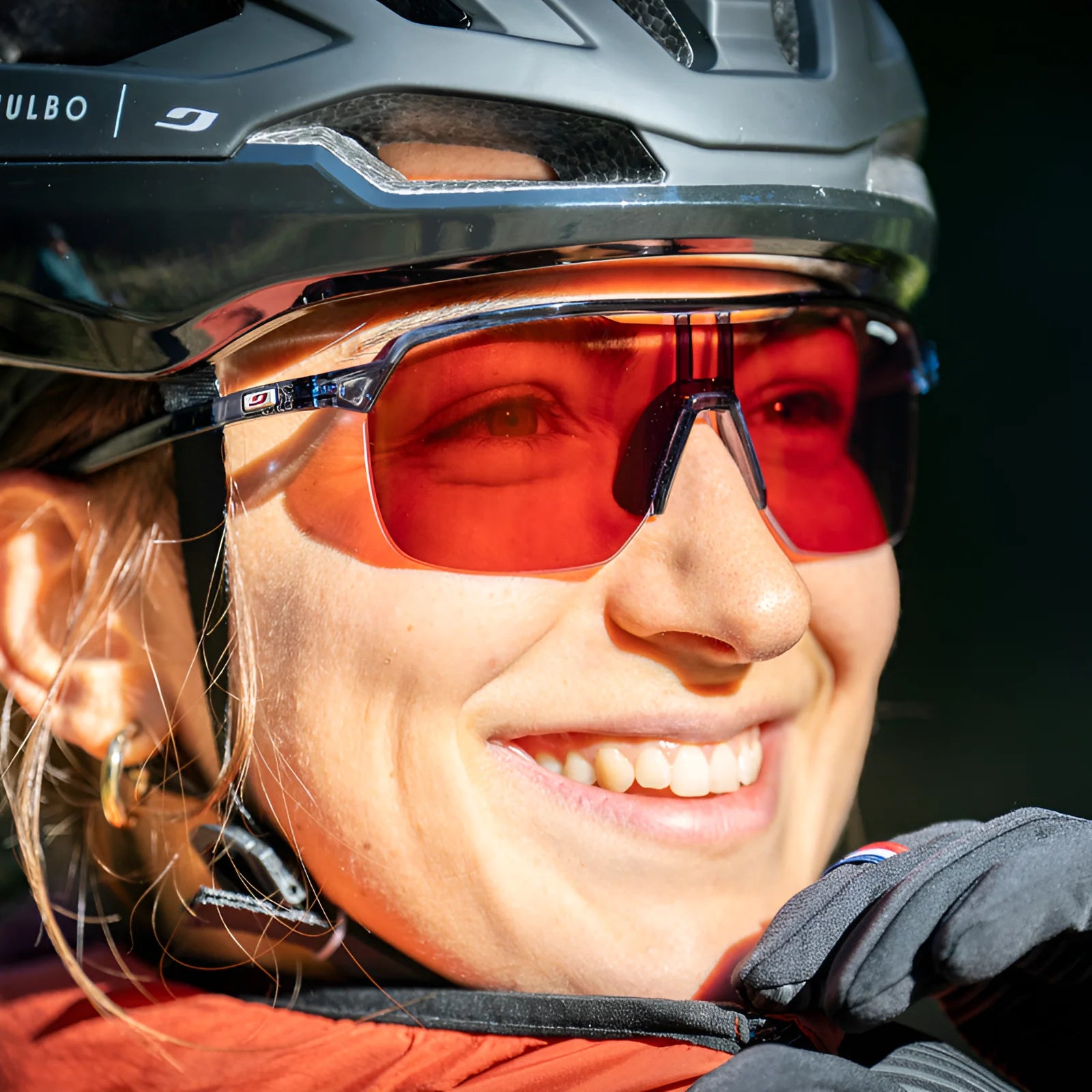 Grinning, this person embraces outdoor adventures wearing a helmet, Julbo Frequency Sunglasses, and gloves.